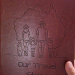 Red leather australian travel journal with embossed design of a family and text 'Our Travel Story' on a wooden surface.