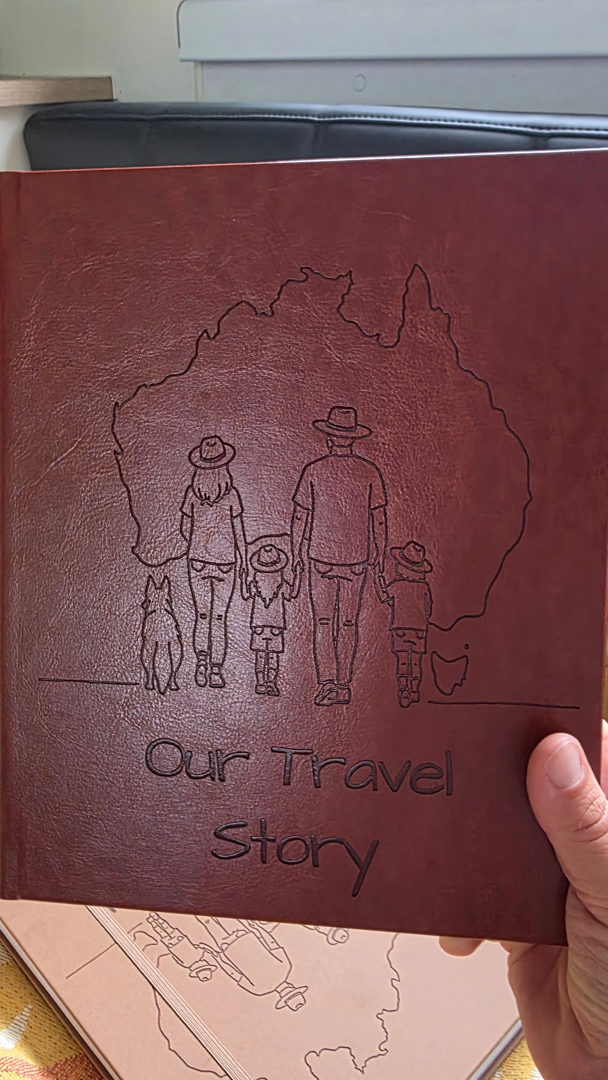 Red leather australian travel journal with embossed design of a family and text 'Our Travel Story' on a wooden surface.