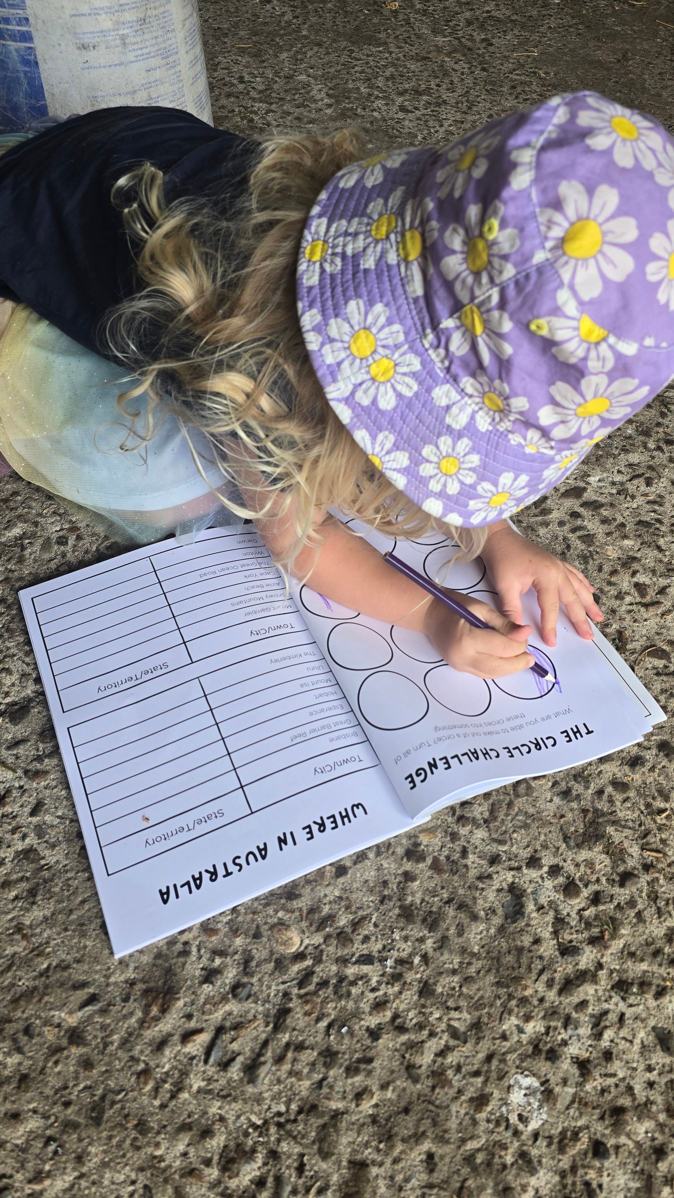 Child wearing a purple floral hat and sitting on the ground with a australian road trip coloring book.