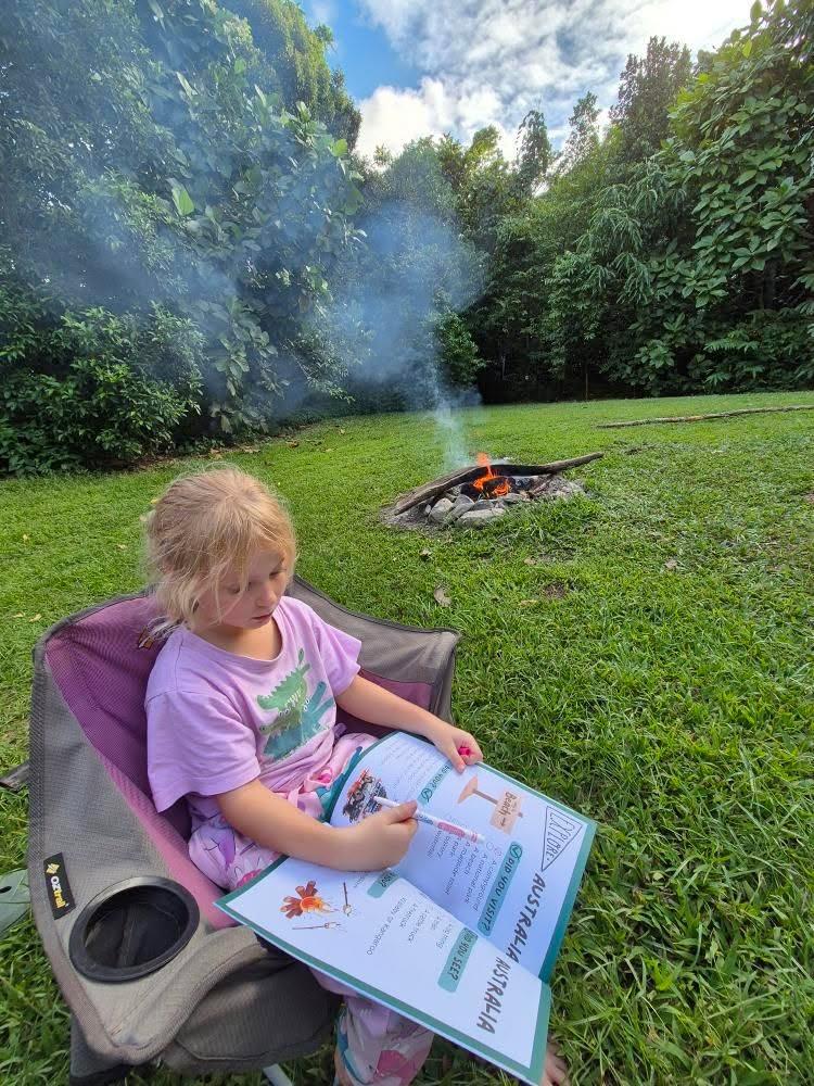 Child sitting in a chair by a campfire in a forest, completing a australian travel journal