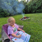 Child sitting in a chair by a campfire in a forest, completing a australian travel journal