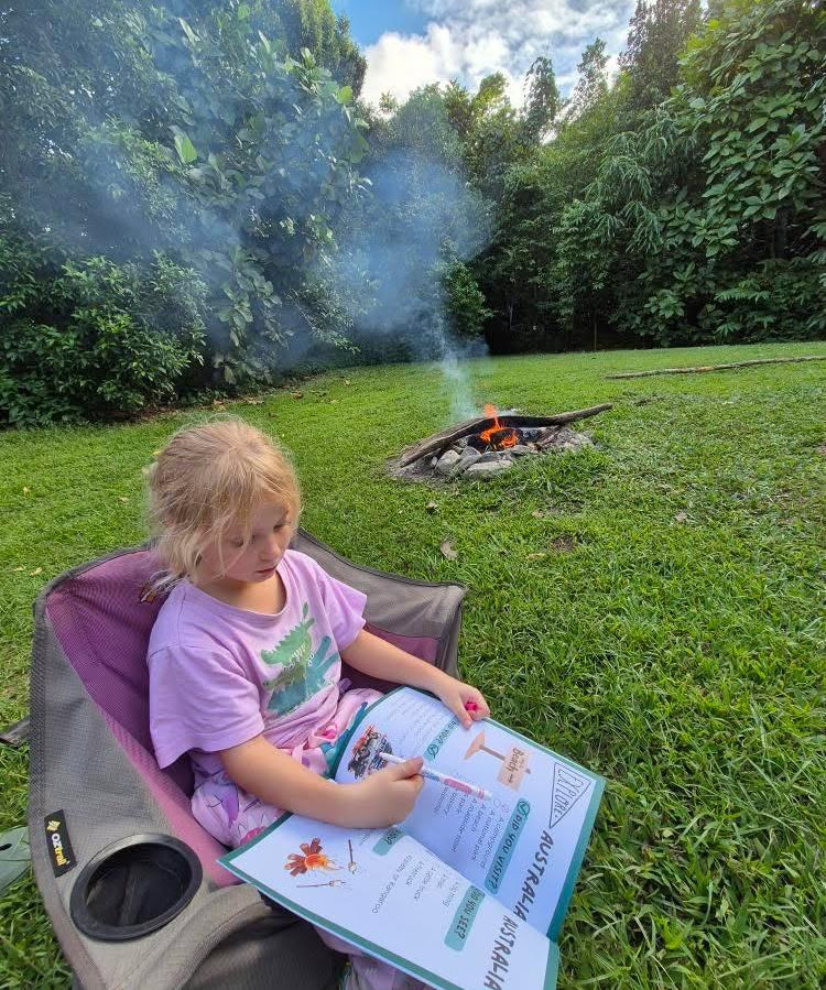 Child sitting in a chair by a campfire in a forest, completing a australian travel journal