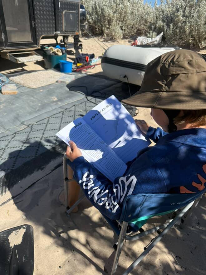 Child sitting outdoors, holding an australian sea life activity book with camping equipment in the background.