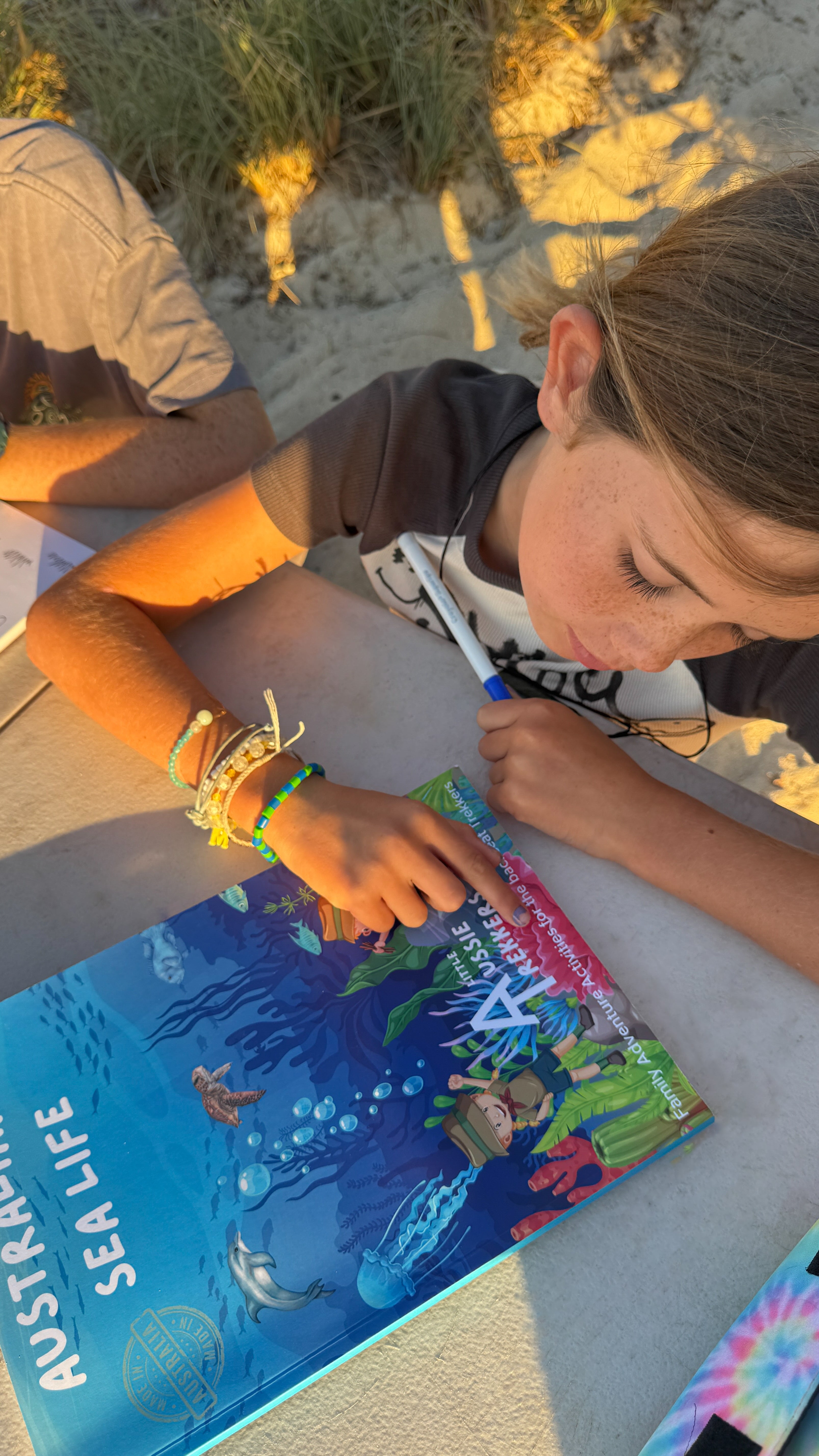 Two children sitting on a beach, one reading a book titled 'Australian Sea Life'.