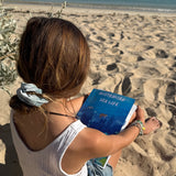 Child sitting on a sandy beach holding a australian sea life activity book, with ocean and clear sky in the background.