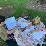 Three children sitting at a table outdoors, engaged in an home schooling australian activity book