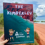 A hand holding a travel journal titled 'THE KIMBERLEY' with an illustration of a little aussie trekker, set against a backdrop of a Kimberley landscape.