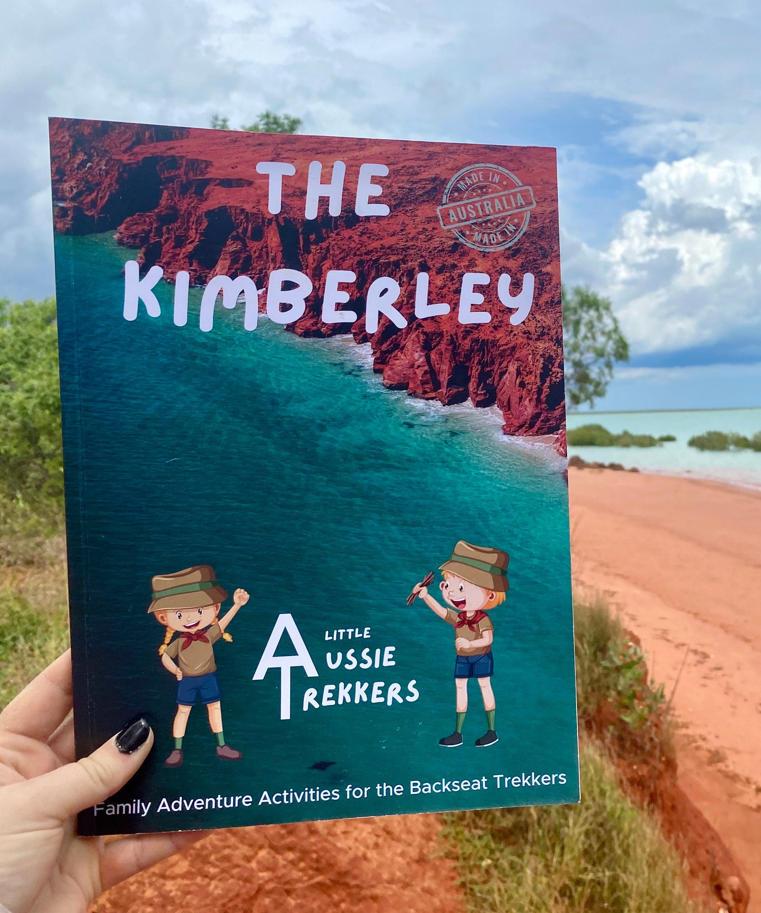 A hand holding a travel journal titled 'THE KIMBERLEY' with an illustration of a little aussie trekker, set against a backdrop of a Kimberley landscape.