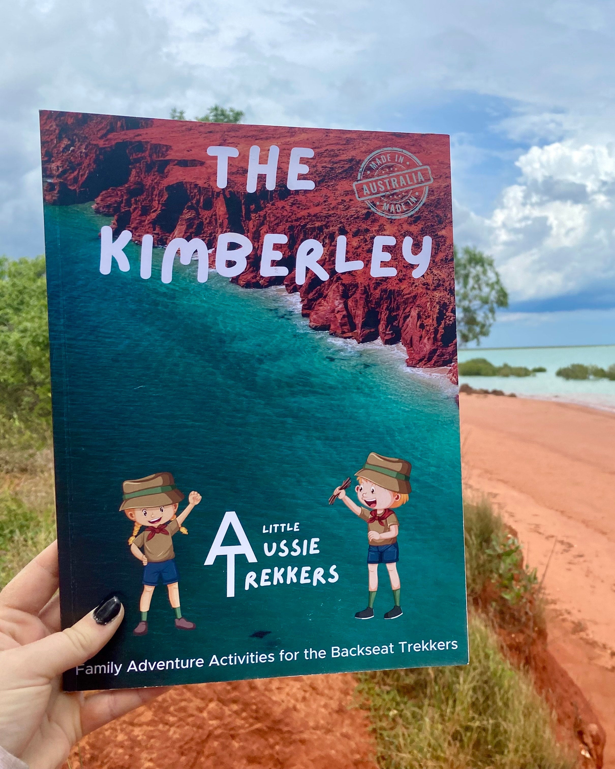 A hand holding a travel journal titled 'THE KIMBERLEY' with an illustration of a little aussie trekker, set against a backdrop of a Kimberley landscape.