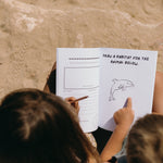 Two children sitting on sand, looking at a book with a drawing of a dolphin and instructions to draw its habitat.