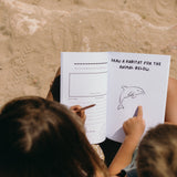 Two children sitting on sand, looking at a book with a drawing of a dolphin and instructions to draw its habitat.