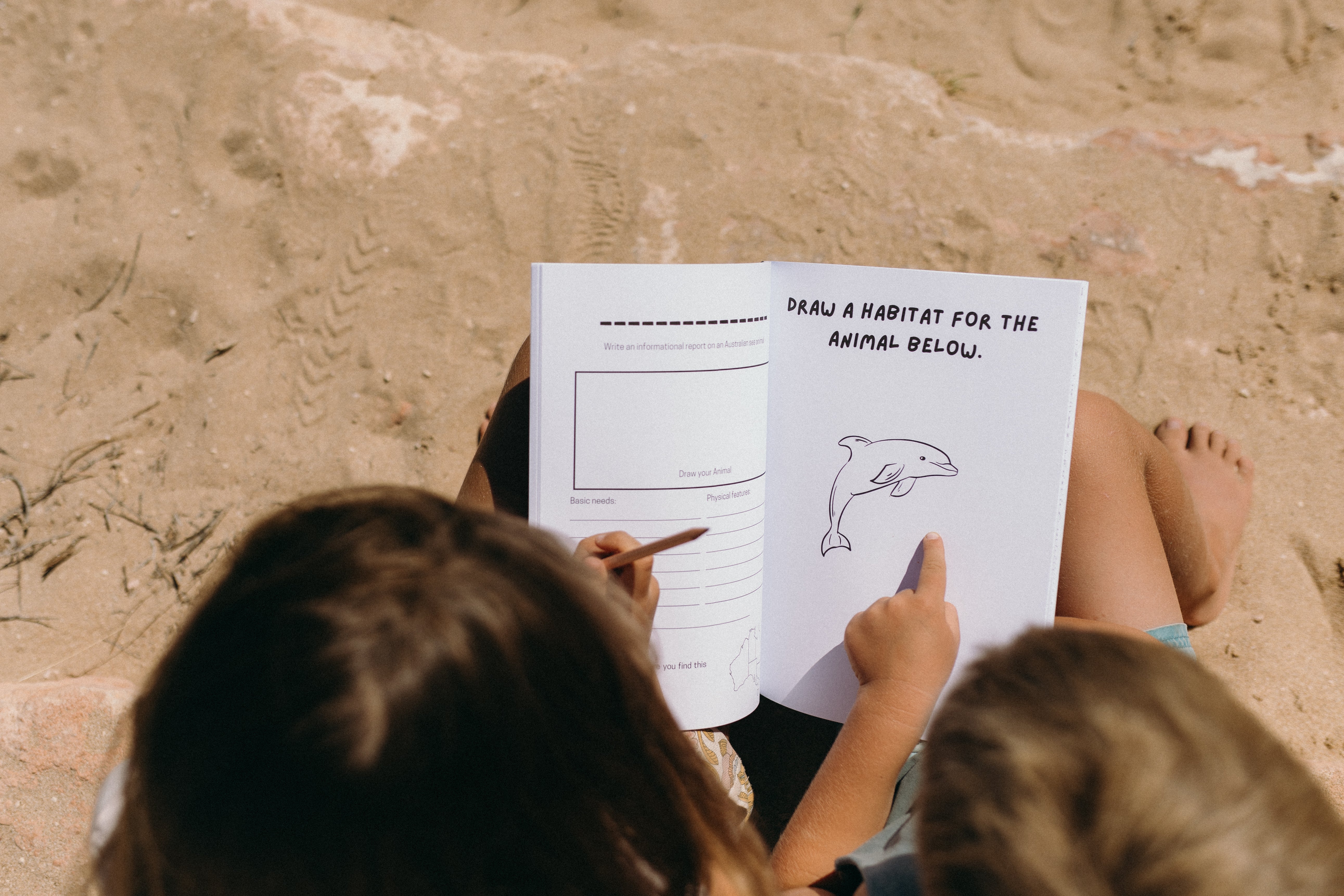 Two children sitting on sand, looking at a book with a drawing of a dolphin and instructions to draw its habitat.