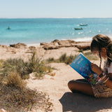 Child reading a australian sea life activity book on a sandy beach with ocean and boats in the background