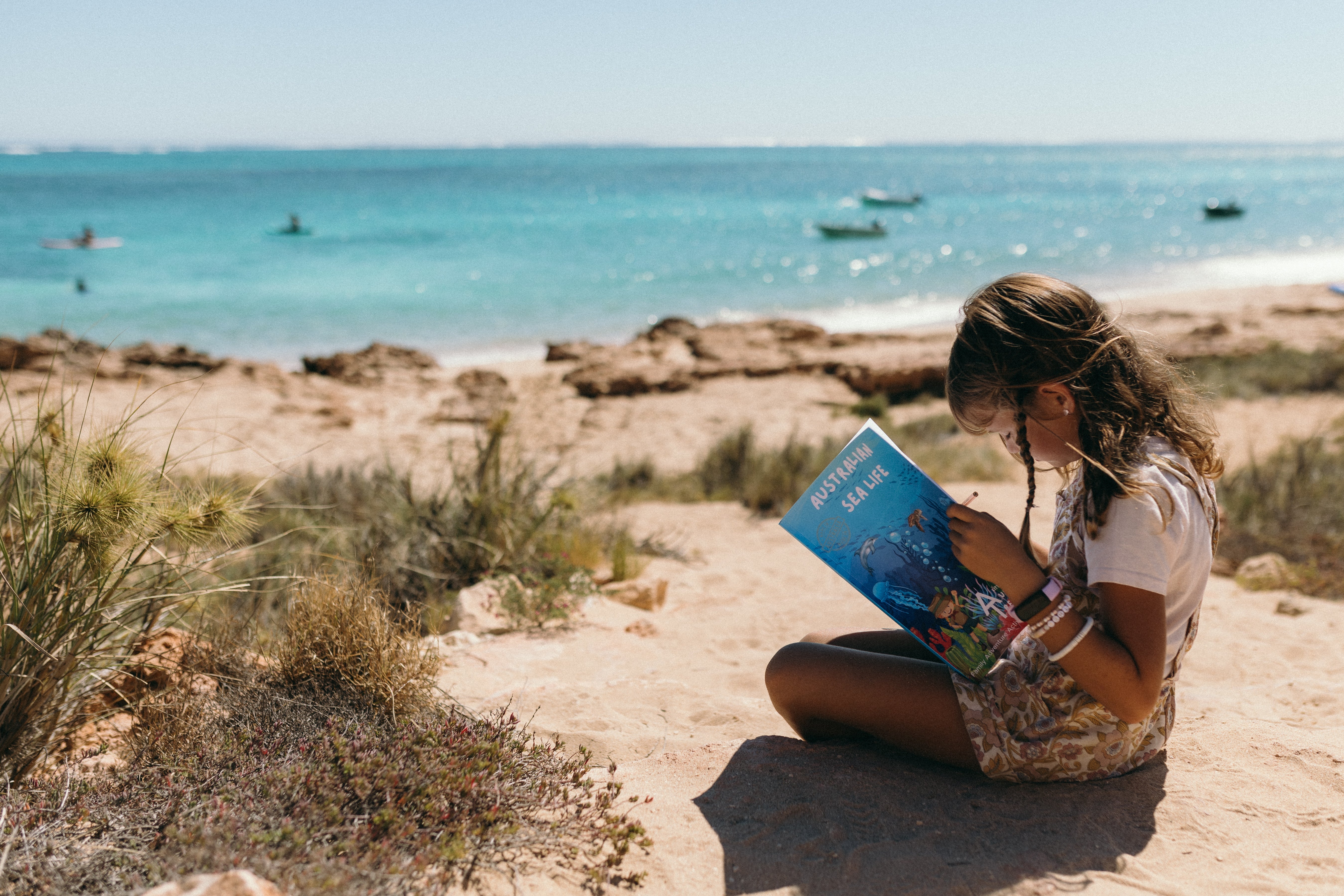 Child reading a australian sea life activity book on a sandy beach with ocean and boats in the background